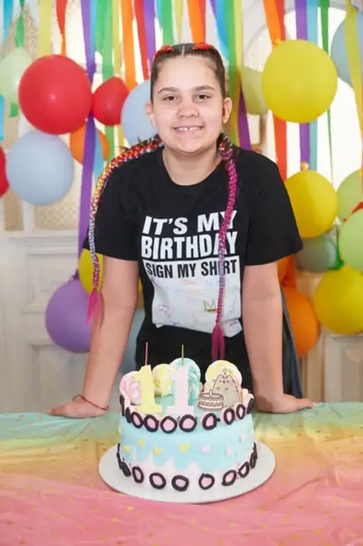 Момиче с цветни плитки и тениска „It's my birthday“ стои зад двуетажна торта пред стена от балони и хартиени ленти.