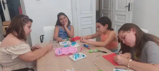 Четири момичета работят с бои, хартия и калъпи около дървена маса.