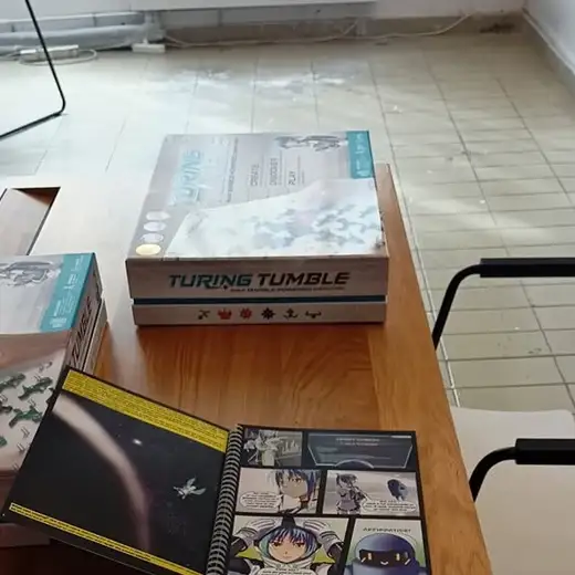 Две кутии на играта Turing Tumble и разгърната книжка с комикс върху дървена маса.