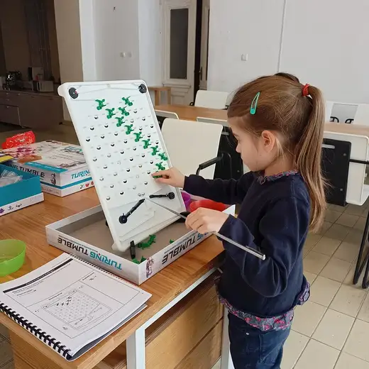 Момиче нарежда зелени детайли по вертикалната дъска на Turing Tumble, до нея кутията и книжка с задачи.
