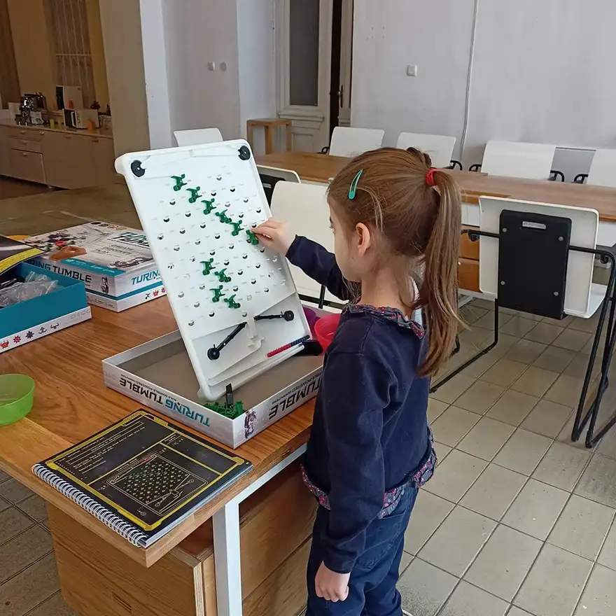 Момиче поставя зелена детайлна част върху вертикална игрална дъска Turing Tumble на маса в зала с плочки и бели столове.