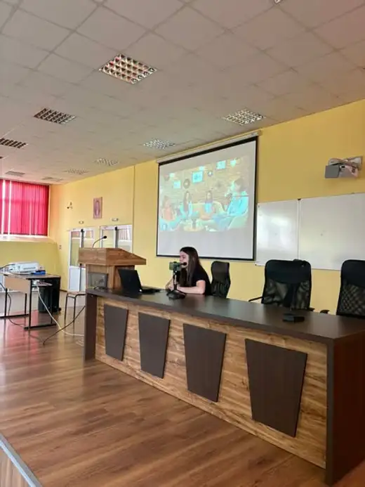 Жена стои зад катедра в зала, а на екрана зад нея е проектирана снимка на деца.