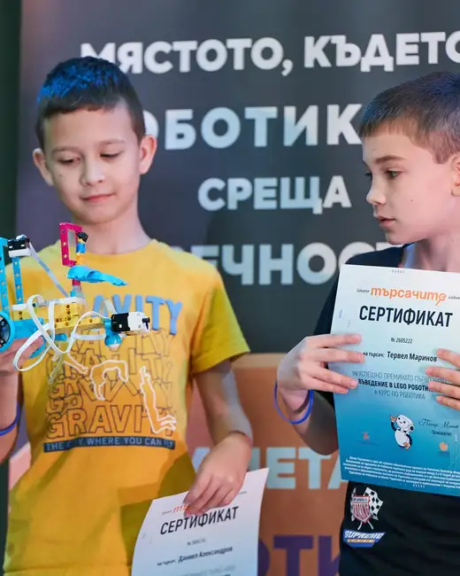 Две момчета пред тъмно пано с надпис за роботиката: едното държи робот от LEGO, другото – сертификат.