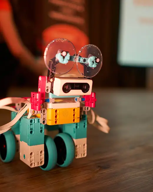 Малък робот от LEGO на сини колела с жълт корпус и две прозрачни кръгли „очи“ върху дървена маса.