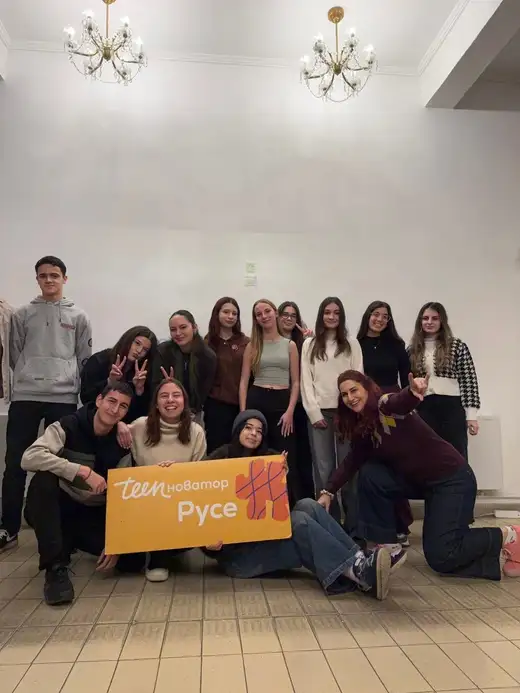 Група младежи в светла зала с полилеи позират около оранжев банер „Teenovator Русе“.