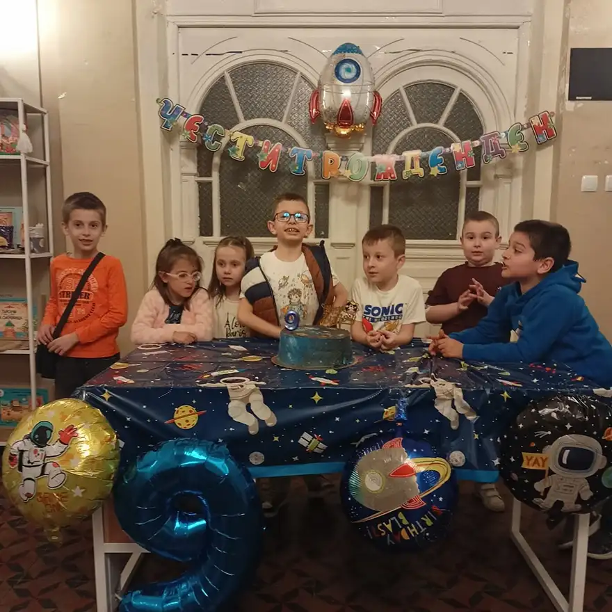 Деца стоят около маса с покривка на тема Космос, върху нея има синя торта със свещичка, а над масата е закачен балон-ракета.