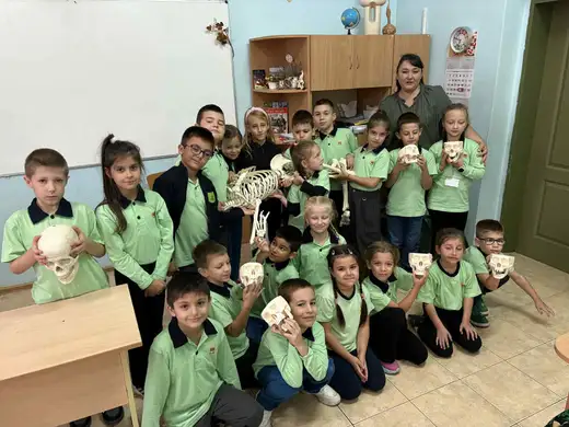 Ученици в зелено се снимат в класната си стая с модел на скелет и черепи в ръце.